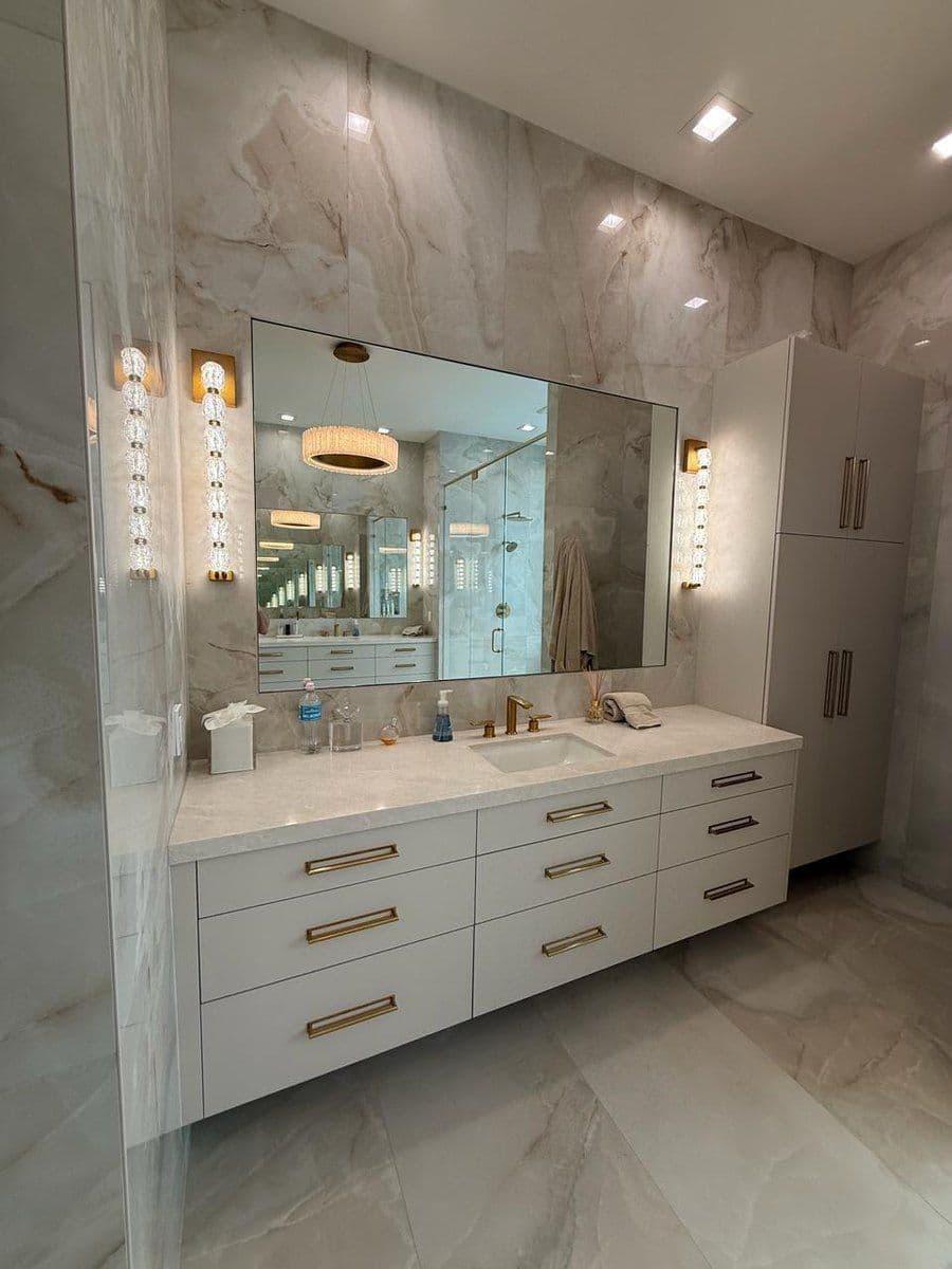 Luxury bathroom remodel with double vanity, large wall mirror, marble-look tile walls, brass hardware, and built-in storage cabinets.